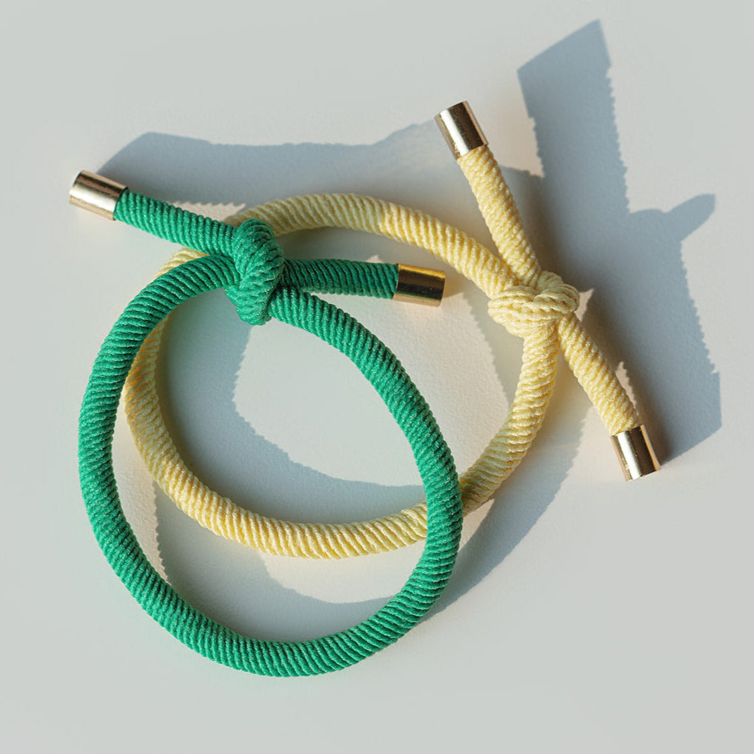 Two braided hair ties, one green and one yellow, on a light gray background.