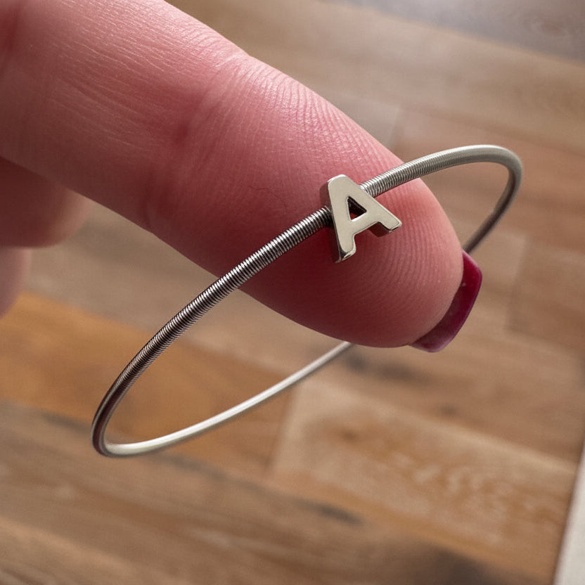 Silver bracelet with a letter 'V' charm held by a finger on a wooden floor.