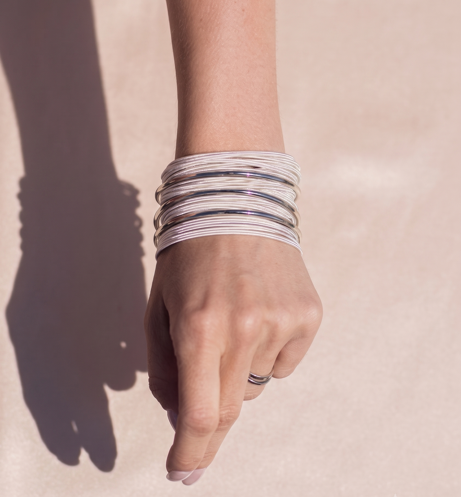 A close-up of a person's hand and forearm wearing a stack of multiple thin metallic bangles on a warm beige background with a strong shadow.