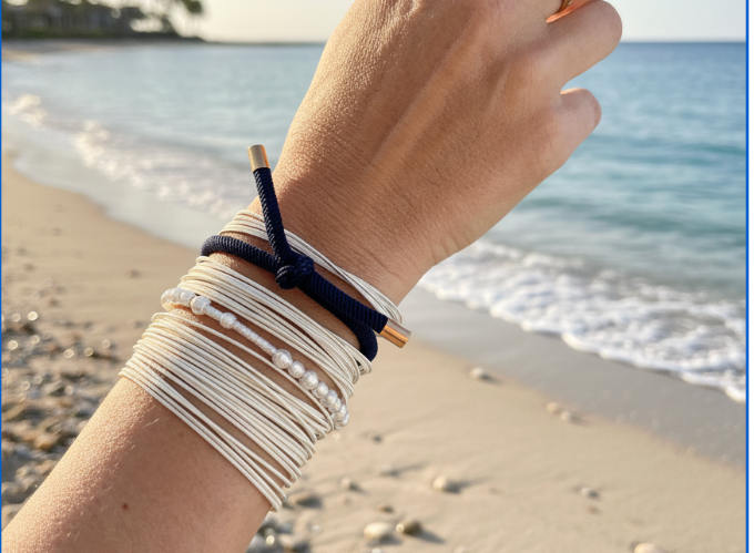 A close-up of a person's wrist at a beach, wearing a stack of white beaded and cord bracelets with one dark navy bracelet and a gold ring on the finger, with ocean waves and sand in the background.
